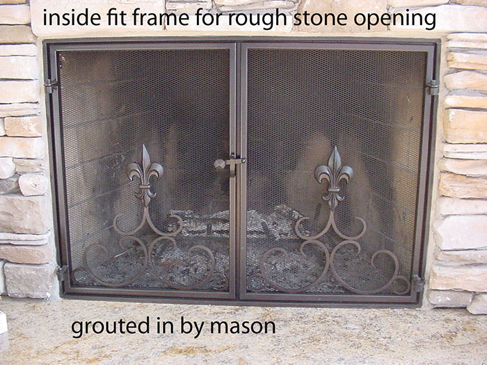 Double door fireplace screen with internal fit frame in a rough stone opening, grouted to fit. Steel has a brown patina and the doors frame a coiled steel and fleur de lis design.