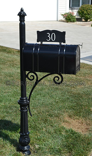 A heavy iron mailbox post with the contemporary bracket standing in grass with a house and driveway just visible in the background.