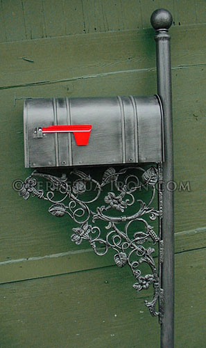 Mailbox post leaning up againsta  green wall ready for installation. This mailbox has the double banded style with avery large fillagreed design.